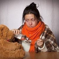 A woman is wrapped up warmly, appearing to have the flu, pouring medicine next to her teddy bear